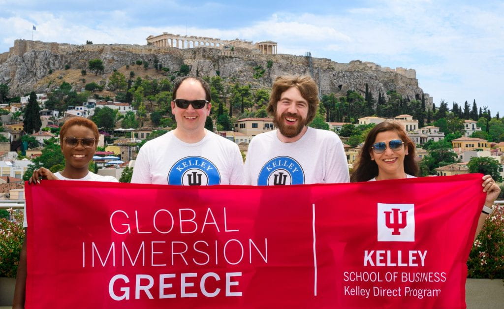 Four Kelley Direct Online MBA students stand in front of the Acropolis in Athens, Greece, holding a flag that says "Global Immersion: Greece."